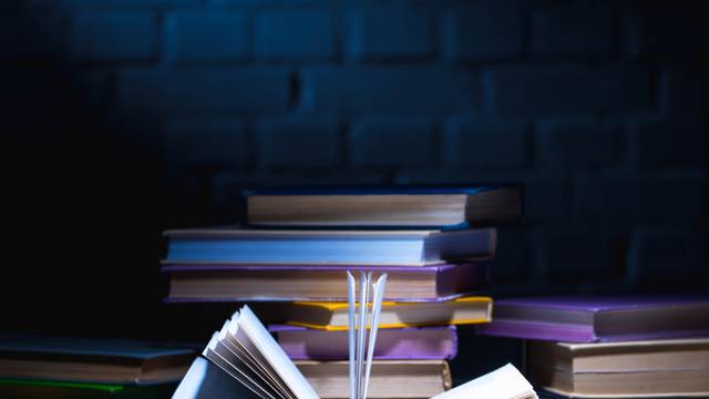 Ein aufgeschlagenes Buch liegt im Vordergrund, dahinter mehrere gestapelte Bücher, beleuchtet in dramatischem Licht vor einer dunklen Ziegelwand