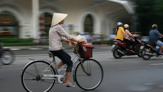 Radfahrerin mit typischem Hut fährt durch vietnamesische Stadt