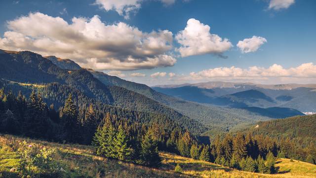 Weite Berglandschaft mit dichten Wäldern und grünen Wiesen im Vordergrund, unter einem Himmel mit weißen Wolken, die lange Schatten auf die Hügel werfen