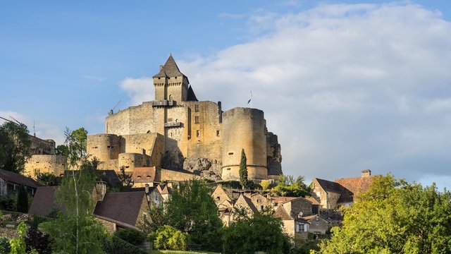 Mittelalterliche Festung Château de Castelnaud in der Dordogne. Die Burg thront auf einem Hügel über einem Dorf mit Steinhäusern und roten Dächern, umgeben von grüner Landschaft. Im Zentrum erhebt sich der mächtige Bergfried mit Spitzdach, flankiert von runden Wehrtürmen.