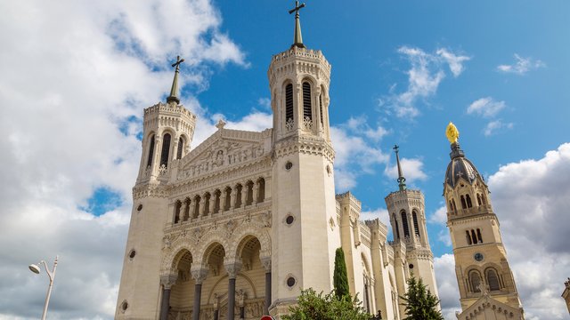 Die Basilika Notre-Dame de Fourvière in Lyon. Ein monumentales weißes Gebäude im neugotischen und byzantinischen Stil mit mehreren Türmen, verzierten Fassaden und einem goldenen Marienstandbild auf einem Turm. Der Himmel ist blau mit einigen Wolken.