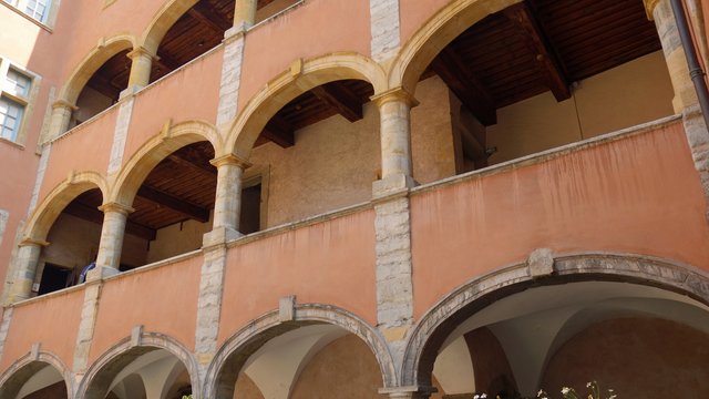 Altbau in der Altstadt von Lyon mit mehrstöckigen Arkadengängen. Die Fassade ist ockerfarben gestrichen, die Bögen und Säulen bestehen aus hellem Stein. Unter den Arkaden wachsen Blumen, die das historische Ambiente ergänzen.