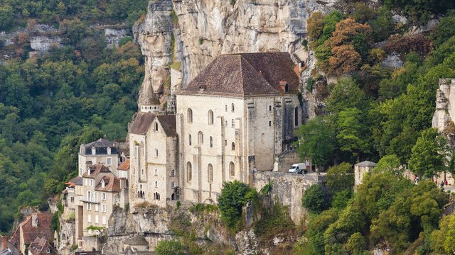 Der Wallfahrtsort Rocamadour in Frankreich. Mehrstöckige mittelalterliche Gebäude und Kirchen sind in die steilen Felswände gebaut. Unterhalb ziehen sich enge Gassen mit Häusern aus hellem Stein und roten Dächern durch das Tal, während oben eine Burganlage mit Kapelle über der Szenerie thront.
