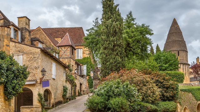 Malerische Gasse in Sarlat-la-Canéda mit historischen Sandsteinhäusern, roten Ziegeldächern und bewachsenen Fassaden. Ein gepflasterter Weg führt bergauf zwischen den Gebäuden hindurch. Rechts im Hintergrund ragt der markante, spitze Turm der Lanterne des Morts über die Bäume.